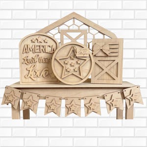 May include: A set of light-coloured wooden patriotic decorations featuring a house-shaped frame, a sign reading America Land that I Love, a circular star plaque with a Stars and Stripes tag, a barn door sign with a bow, and a star-themed bunting banner.