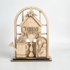 May include: Wooden spring-themed decoration featuring a birdhouse, watering can with flowers, and tulips. The words "HELLO SPRING" are prominently displayed. The design includes a butterfly and is set against a window-like frame.