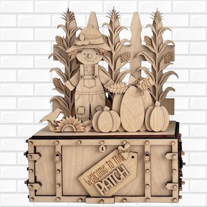 May include: A laser-cut wooden display featuring a scarecrow, corn stalks, and pumpkins standing before a fence. A tag attached to a pumpkin reads Hello FALL, and a larger tag on the base reads Welcome to the PATCH. The piece is light brown.