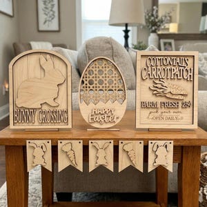 Könnte beinhalten: Holz-Osterdekorationen, darunter ein Schild mit "Bunny Crossing" und einer Kaninchensilhouette, eine eiförmige Dekoration mit "Happy Easter" und ein Schild mit "Cottontail's Carrotpatch". Ein Banner mit Hasen- und Karottenausschnitten ist ebenfalls zu sehen.