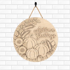 Könnte beinhalten: Ein rundes Wandschild aus hellem Holz mit dem lasergeschnittenen Schriftzug Autumn Greetings. Das Design zeigt geschichtete Holzmotive von drei Kürbissen, zwei Sonnenblumen, Weizenähren, Blättern und Eicheln, aufgehängt an einem beigen Seil.