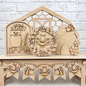 Könnte beinhalten: Ein helles Holz-Dekorationsset für Etagèren mit einem Schild mit der Aufschrift LOCAL HONEY FOR SALE organic raw natural, einem Kranz mit Biene, einem Bienenstock mit Biene und Blumen sowie einem Banner mit fünf einzelnen Bienenwimpeln.