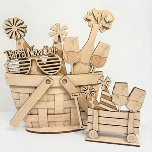 May include: A wooden New Year's Eve-themed basket and cart set. The basket contains champagne, glasses, party hats, and sunglasses with the text "Happy New Year." The cart has glasses and party hats. All items are made of light-colored wood.