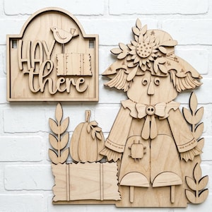 Puede incluir: Un conjunto de decoración de pared de madera clara que incluye un espantapájaros con sombrero de girasol, una calabaza y un letrero rectangular con el texto "HAY there" junto a un pequeño pájaro sobre una paca de heno, todo en madera cortada.