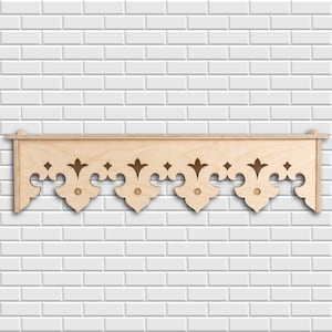 May include: A light-colored wooden shelf featuring a decorative scalloped edge with cut-out fleur-de-lis and circular patterns. The shelf has a flat top surface and a symmetrical, intricate design along the front panel, crafted from natural wood.