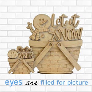 May include: Two light-colored wooden basket-shaped decorations. The larger basket features a snowman with a scarf and the text Let it SNOW. The smaller basket features a smiling face and the text Winter WISHES. Both have woven patterns.