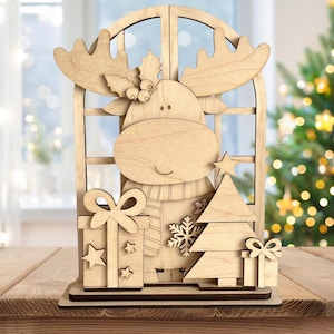 May include: A wooden Christmas decoration featuring a moose in a window frame. The moose wears a scarf and is surrounded by a Christmas tree, presents, and a snowflake. The decoration is made of light-colored wood and stands on a wooden surface.