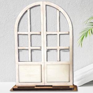 Op de afbeelding: Een decoratief houten raamkozijn met een gewelfde bovenkant en verdeelde ruiten. Het frame is geschilderd in een verweerde witte afwerking en staat op een bruine basis. Geschikt voor interieurdecoratie.