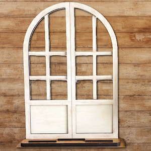 May include: A white, arched window frame with a distressed finish. The frame is divided into multiple panes and has a small shelf at the bottom. The window is made of wood and has a rustic, farmhouse style.