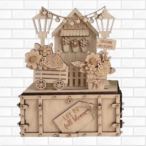 May include: A laser-cut wooden display featuring a flower shop scene with a cart, potted flowers, and string lights. The base is a rectangular box with a hanging tag that reads "LIFE IN full bloom" and signs saying "FRESH FLOWERS" and "SUNSHINE and BLOOMS".