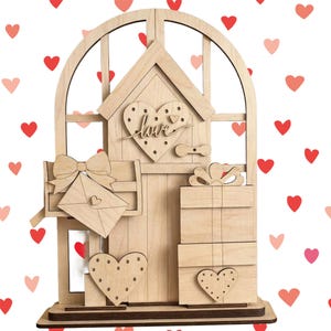 May include: A wooden Valentine's Day ornament in the shape of a house, with a heart-shaped door and window. The word "love" is scripted on the door. It includes a mailbox with an envelope, gift boxes, and heart-shaped embellishments.