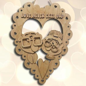 Op de afbeelding: Houten hartvormige wanddecoratie met een ouder echtpaar met een bril. De tekst "LOVE NEVER GETS OLD" is bovenaan gegraveerd. De rand heeft hartvormige uitsparingen. Natuurlijke houten afwerking.