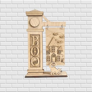 Puede incluir: Un letrero decorativo de madera clara con un poste vertical que tiene la palabra BOO recortada, donde la letra O contiene una araña y una telaraña. Un panel rectangular cuelga del poste, mostrando una casa encantada, murciélagos y un gato.
