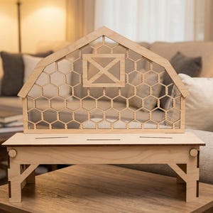 May include: A wooden decorative barn-shaped structure with a chicken wire design. The barn sits on a matching wooden table. The barn has a gable roof and a window with an X-shaped design. The table has angled legs.