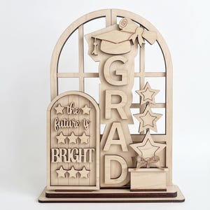 May include: Wooden graduation decoration featuring the word "GRAD" and the phrase "the future is BRIGHT." Includes a graduation cap, diploma, and star accents. The design is made of light-colored wood and is suitable for celebrating a graduation.