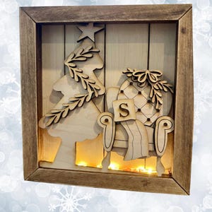 Könnte beinhalten: Weihnachtliche Holz-Schattenbox mit Baum- und Sessel-Design. Der Baum hat einen Stern, und der Sessel hat ein "S" auf einem Kissen. Der braune Rahmen umgibt einen warm beleuchteten Innenraum.