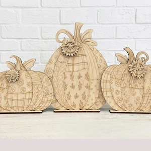 May include: Three light-colored wooden pumpkin cutouts of varying sizes, each featuring intricate laser-engraved patterns including flowers, leaves, and plaid designs. Each pumpkin is topped with a small wooden sunflower embellishment.