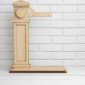 Puede incluir: Un soporte de exhibición de madera clara que presenta un pilar vertical con una base cuadrada y un brazo horizontal que se extiende desde la parte superior. La estructura incluye un detalle circular en el pilar y una base rectangular plana.