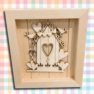 May include: Wooden wall art featuring a heart-shaped birdhouse design. The artwork includes laser-cut birds, hearts, and floral accents. The piece is framed in a light-colored wood, creating a charming, rustic aesthetic.