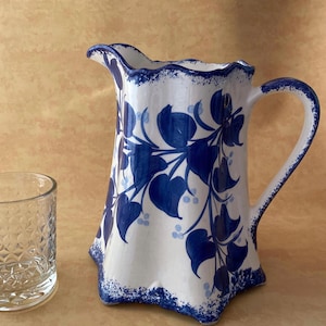 May include: White ceramic pitcher with a blue floral design. The pitcher has a scalloped rim and handle. A clear glass with a textured pattern is to the left of the pitcher.