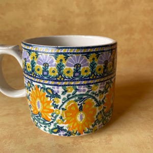 May include: A ceramic mug with a white handle and rim, featuring a floral pattern in orange, yellow, green, and purple on a white background. The mug has a cylindrical shape and a curved handle.