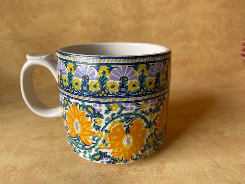 May include: A ceramic mug with a white handle and rim, featuring a floral pattern in orange, yellow, green, and purple on a white background. The mug has a cylindrical shape and a curved handle.