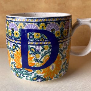 May include: A white ceramic mug with a black handle and the text "But First, Coffee" in a bold, black font. The mug is filled with a dark liquid, likely coffee, and sits on a light-colored surface.