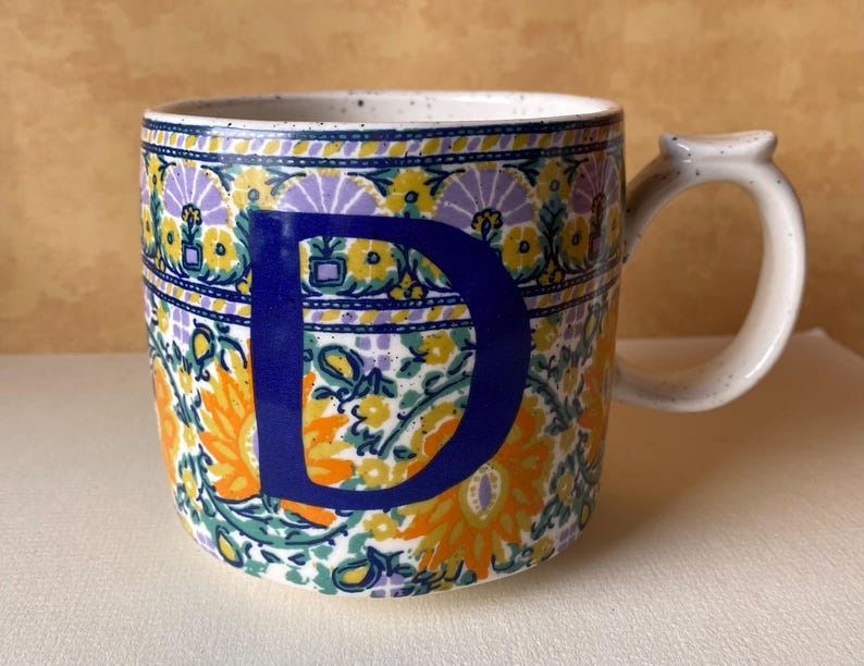 May include: A white ceramic mug with a black handle and the text "But First, Coffee" in a bold, black font. The mug is filled with a dark liquid, likely coffee, and sits on a light-colored surface.
