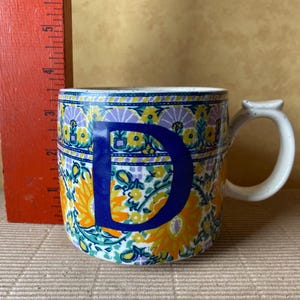 May include: A ceramic mug with a floral pattern in yellow, orange, and teal, and a large blue letter "D". The mug has a white handle and a speckled rim. A red ruler is visible in the background.