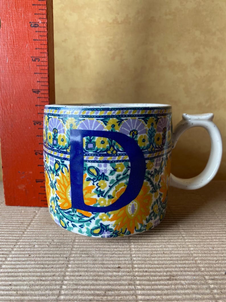 May include: A ceramic mug with a floral pattern in yellow, orange, and teal, and a large blue letter "D". The mug has a white handle and a speckled rim. A red ruler is visible in the background.