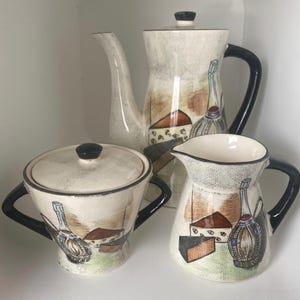May include: A vintage ceramic tea set featuring a cream-colored background with black handles and accents. The set includes a teapot, sugar bowl, and creamer, each decorated with a wine bottle, cheese, and landscape design.