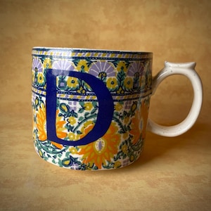 May include: A ceramic mug with a white handle and a floral pattern in yellow, orange, purple, and green. A large blue letter "D" is prominently displayed on the front of the mug.