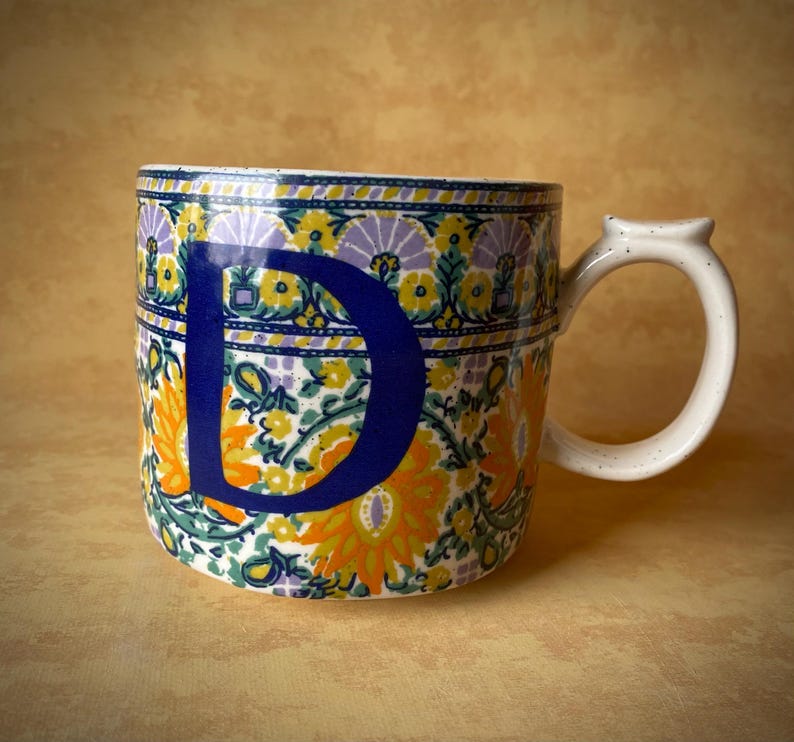 May include: A ceramic mug with a white handle and a floral pattern in yellow, orange, purple, and green. A large blue letter "D" is prominently displayed on the front of the mug.