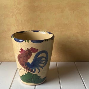 May include: A cream-colored ceramic planter featuring a hand-painted rooster design with brown, blue, and green details. The rim is decorated with a series of blue circular patterns. The vessel has a tapered cylindrical shape and a smooth finish.