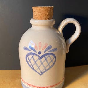 May include: A small, off-white ceramic jug with a cork stopper. The jug features a blue and pink heart design and a handle. A blue and red band decorates the base. The jug is approximately 5 inches tall.