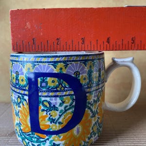 May include: A ceramic mug with a white handle and a large blue letter "D". The mug is decorated with a floral pattern in yellow, orange, purple, and teal. The mug is approximately 4 inches tall.