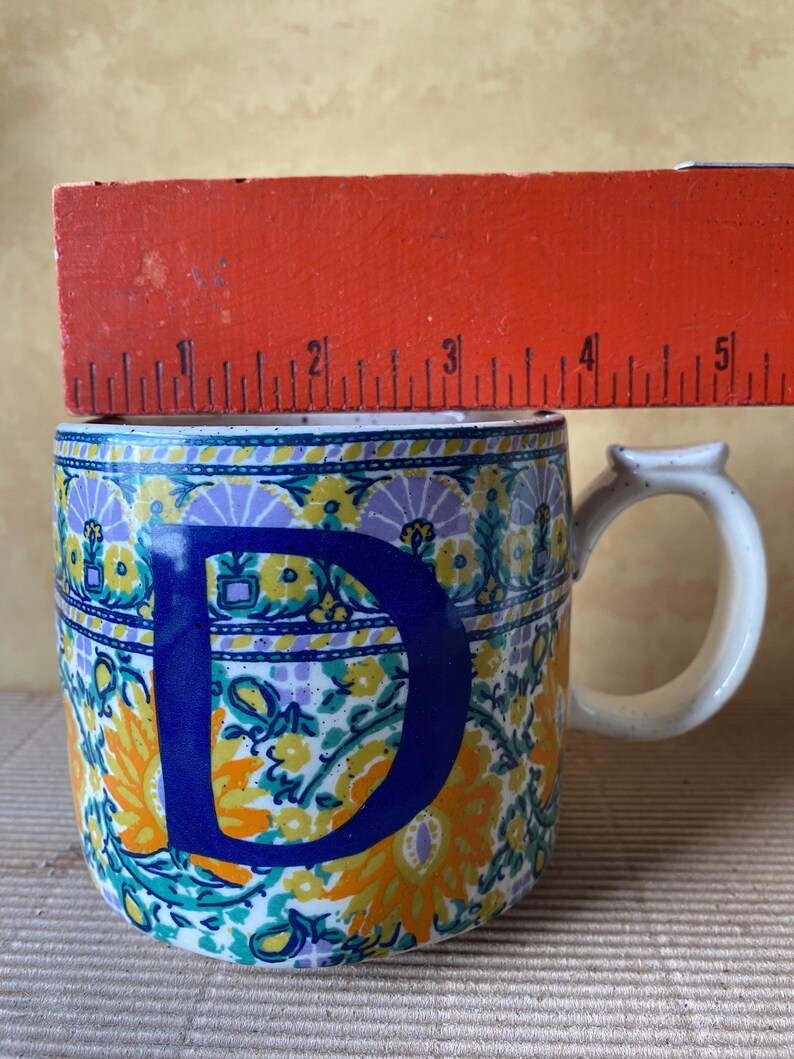 May include: A ceramic mug with a white handle and a large blue letter "D". The mug is decorated with a floral pattern in yellow, orange, purple, and teal. The mug is approximately 4 inches tall.