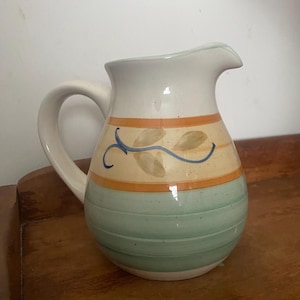 May include: A ceramic pitcher with a cream-colored body, a curved handle, and a spout. The pitcher features a band of orange and yellow with leaf designs and a green base. The pitcher is sitting on a wooden surface.