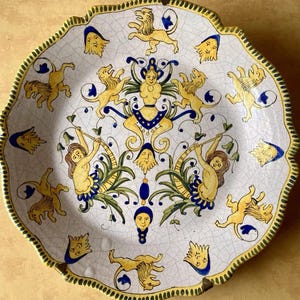May include: A round ceramic plate with a scalloped edge featuring a crackled white glaze. The surface is decorated with yellow lions, blue and yellow stylized faces, and two central figures with long hair surrounded by green foliage and blue accents.