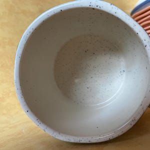 May include: A speckled white ceramic mug with a dark blue rim and handle. The interior of the mug is white with small black speckles. A stack of orange and blue ceramic bowls is visible in the background.