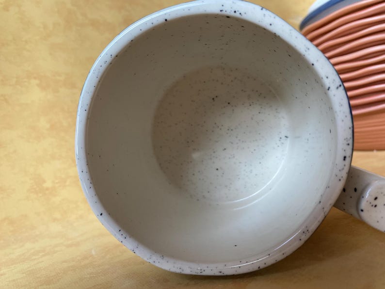 May include: A speckled white ceramic mug with a dark blue rim and handle. The interior of the mug is white with small black speckles. A stack of orange and blue ceramic bowls is visible in the background.