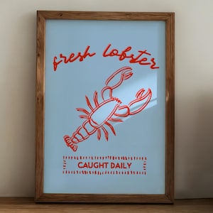 Op de afbeelding: Een ingelijste poster met een lichtblauwe achtergrond toont een rode kreeftenillustratie en de tekst "fresh lobster" en "CAUGHT DAILY". De poster staat op een houten plank met een koffiemachine en keukengerei.