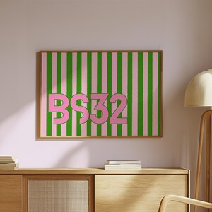 Puede incluir: Una obra de arte enmarcada con un fondo de rayas rosas y verdes y el texto "BS32" en rosa. La obra de arte se exhibe en un mueble de madera clara.