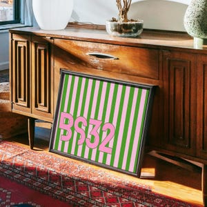 Puede incluir: Una obra de arte enmarcada con un fondo a rayas rosas y verdes y el texto "BS32" en rosa. La obra est&aacute; apoyada en un mueble de madera. La est&eacute;tica general es retro y colorida.