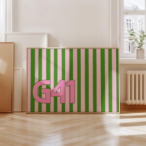 Puede incluir: Una obra de arte enmarcada con un fondo de rayas rosas y verdes y el texto "G41" en rosa. La obra de arte est&aacute; en un marco de madera y se exhibe contra una pared blanca.
