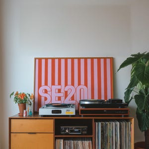 Puede incluir: Un mueble de madera con dos tocadiscos, una obra de arte enmarcada con el texto "SE20" y una colecci&oacute;n de discos de vinilo. La obra presenta rayas verticales en naranja y rosa. Una planta en maceta est&aacute; a la izquierda y una gran planta verde a la derecha.