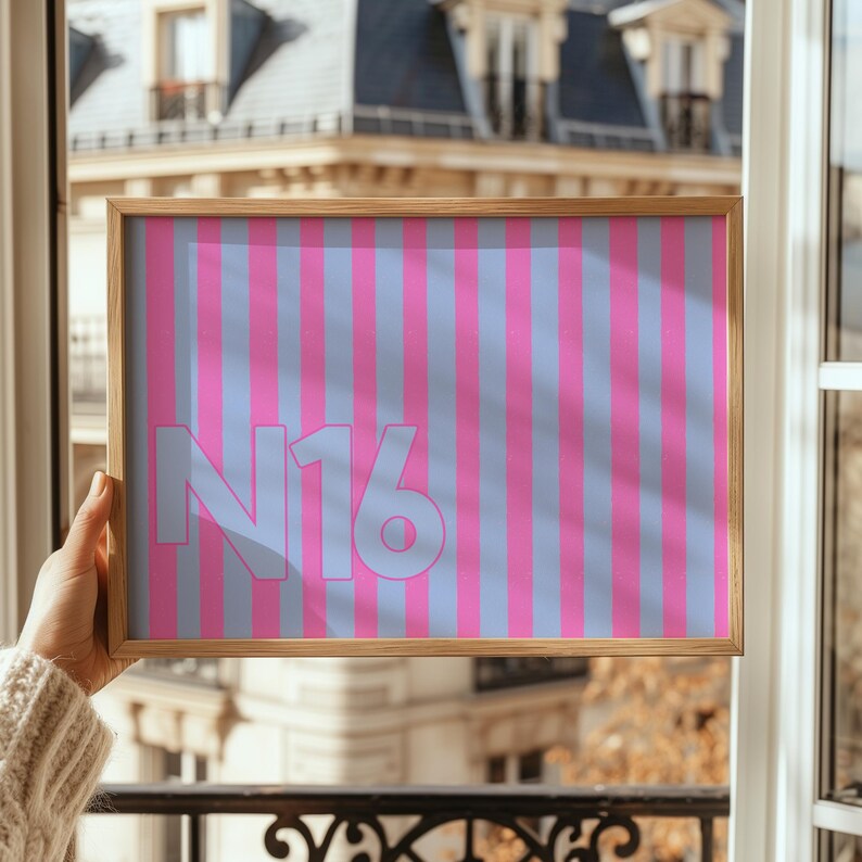 Puede incluir: Un cuadro enmarcado con un patr&oacute;n de rayas rosas y celeste. El cuadro presenta el texto "N16" en celeste. El marco es de madera clara. La obra de arte se sostiene frente a una ventana.