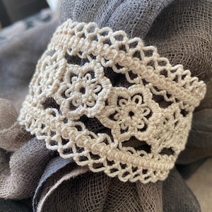 May include: A close-up of a beige crocheted lace trim with a floral pattern. The lace is on top of a brown burlap fabric.