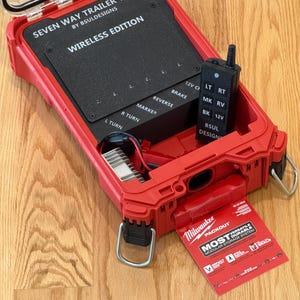 May include: A red trailer tester kit with a clear lid, labeled "SEVEN WAY TRAILER TESTER WIRELESS EDITION." Includes a black control unit and a red Milwaukee Packout label. The kit is open, revealing internal components.
