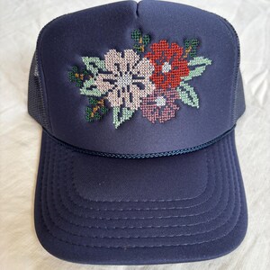 May include: Navy blue trucker hat featuring a floral cross-stitch design. The embroidered flowers include white, red, and pink blooms with green leaves. The hat has a mesh back and a curved peak.